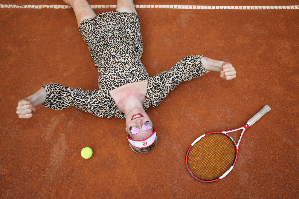 Angela liegt auf dem Boden des Tennisplatzes. Sie trägt ein kurzes Tenniskleid mit einer Sonnenbrille mit Tigerprint und einem Tennishut. Neben ihr liegen ein Tennisschläger und ein Tennisball. Angela ballt ihre Hände zu Fäusten, eine gewinnende Geste. Sie lächelt.